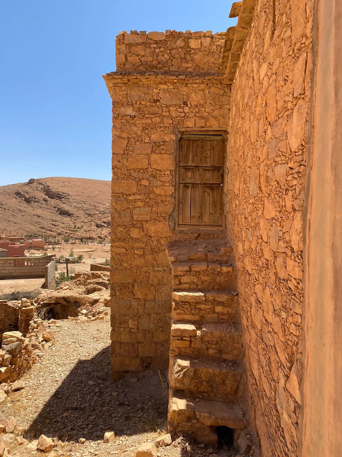 درج من الحجر يصعد بمحاذاة بيت حتى باب من الخشب، وخلفه الجبل وبيوت اليوم.