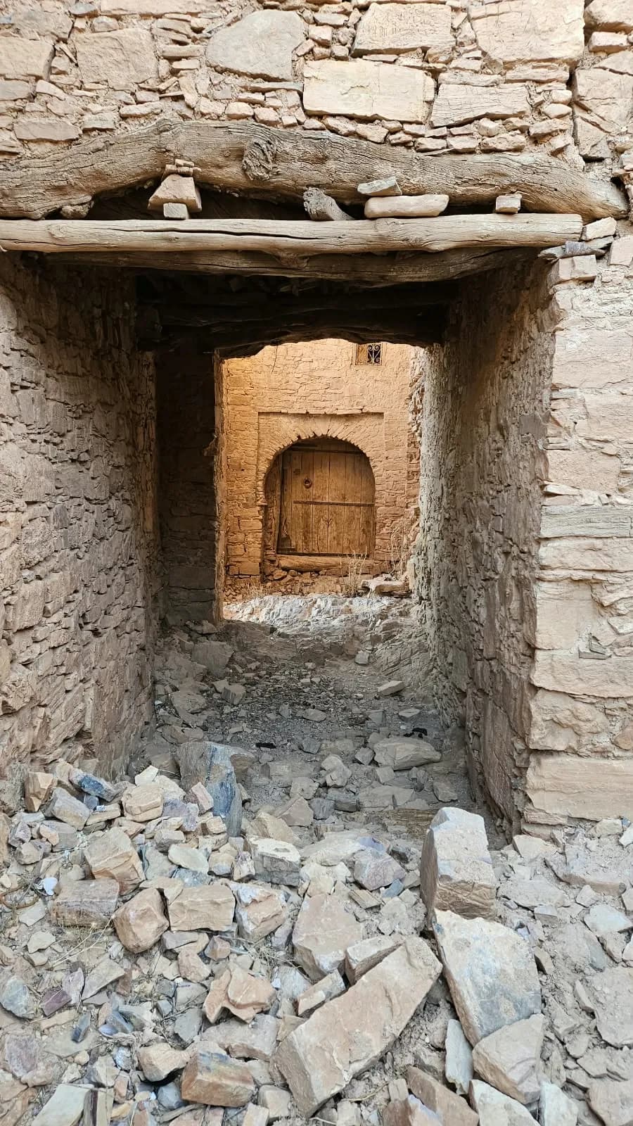 ممرّ مسقوف تحت عارضتين من جذوع الشجر بين جدارين من الحجر، وفي آخره باب خشبي مقوّس في الضوء، فوقه نافذة صغيرة بشبكة حديدية، والحجارة المتساقطة تغطّي الأرض.