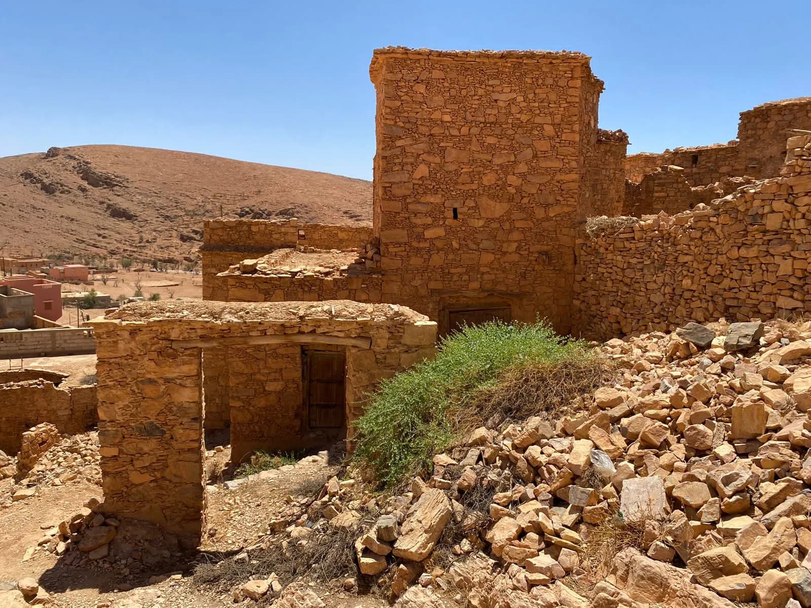 بناية عالية من الحجر وجدرانها، وشجيرة خضراء وسط الحجارة المتهدّمة، وفي البعيد إلى اليسار بيوت اليوم عند سفح الجبل.