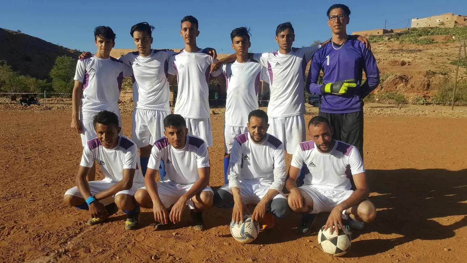 Une équipe en maillot blanc pose sur le terrain de terre, avec son gardien en violet.