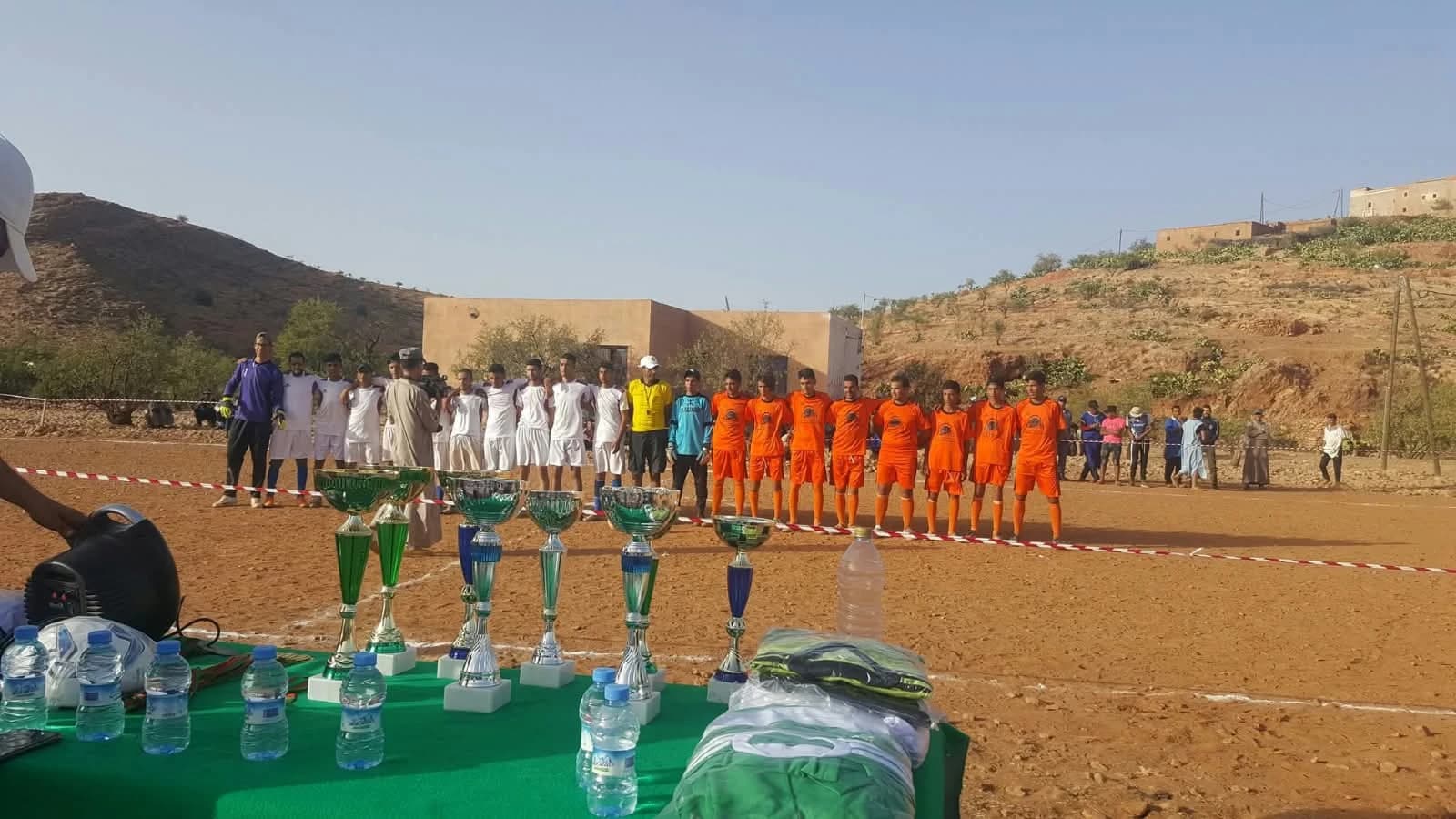 الفريقان مصطفّان في الملعب قبل مباراة، أحدهما بالأبيض والآخر بالبرتقالي، والحكم في الوسط، والكؤوس في المقدمة.